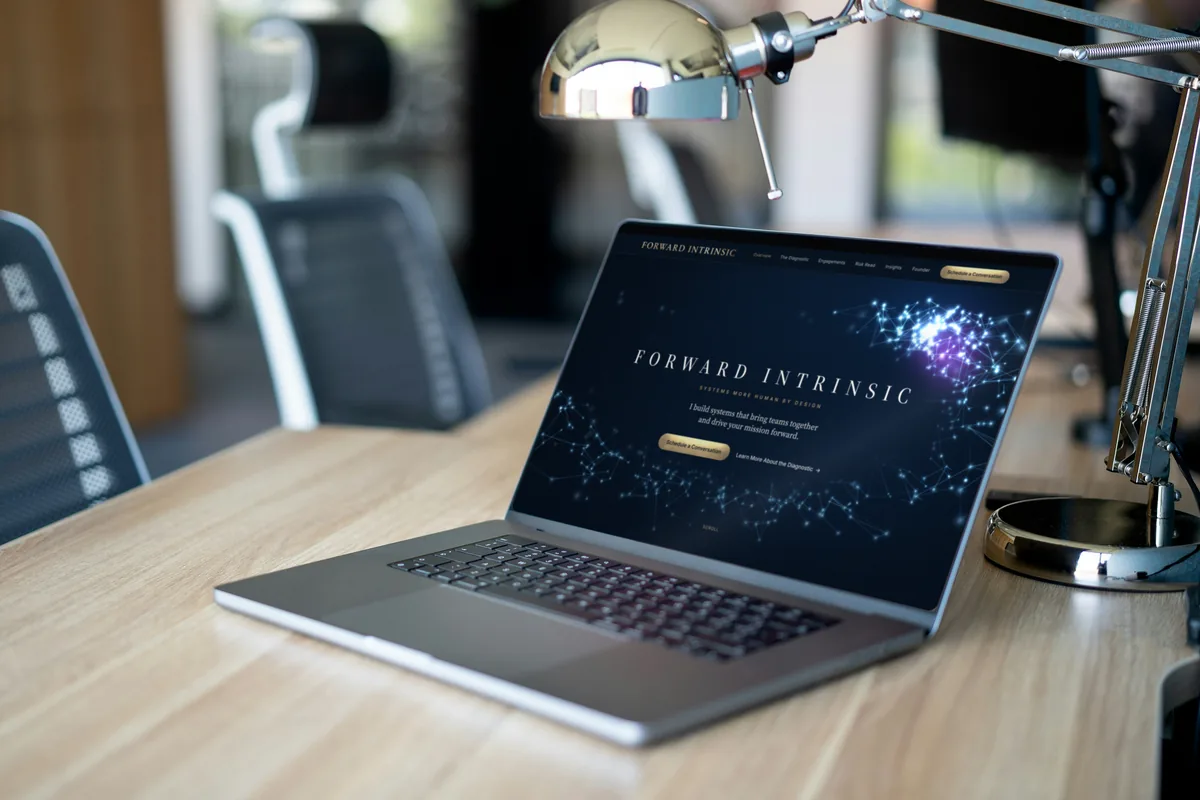 forwardintrinsic.com on a laptop on a wooden desk under a desk lamp